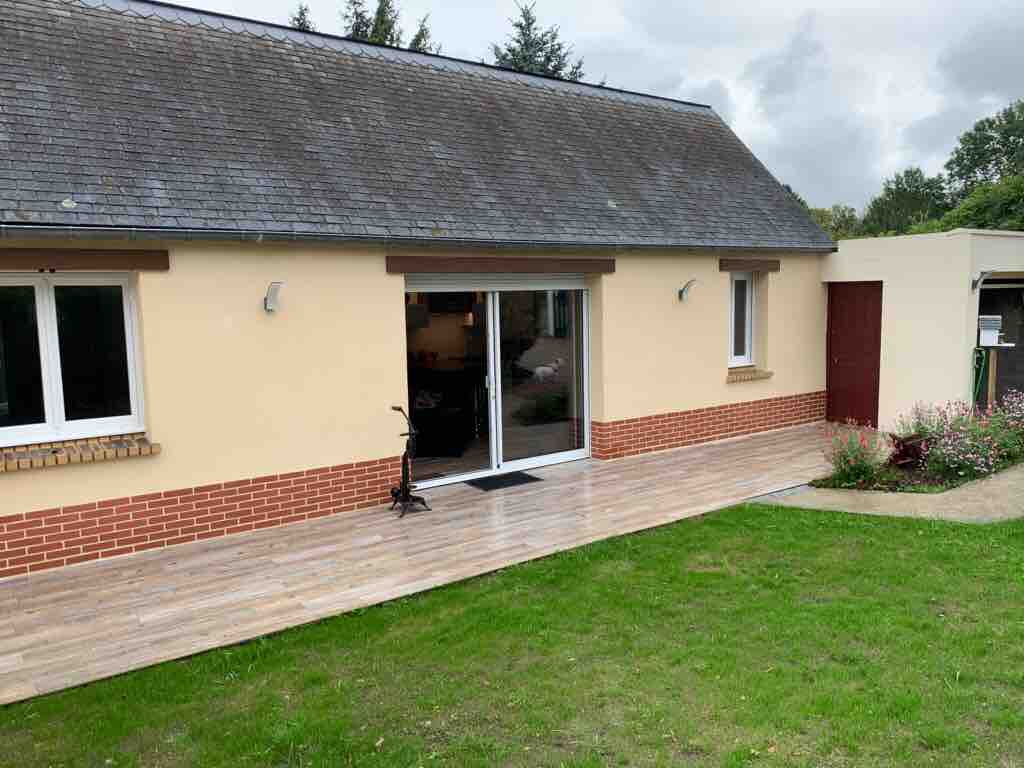 A single-story house features a welcoming exterior with a smooth wooden deck leading to large glass doors. A lush green lawn surrounds the home, complemented by flower beds. Window and door details are highlighted, enhancing the inviting atmosphere of the property.