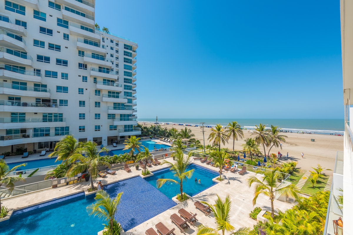An expansive view reveals multiple swimming pools surrounded by palm trees, with lounge chairs positioned for relaxation. Beyond the pools, a golden sandy beach stretches towards the horizon under a clear blue sky.