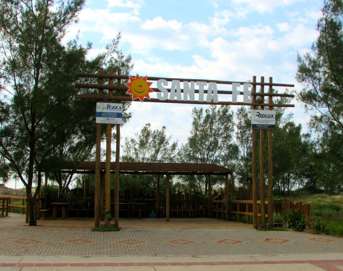 A welcoming entrance to Santa Fe is featured, marked by a large wooden structure with a bright sun emblem. The area is framed by lush greenery, while shaded seating is provided underneath the canopy, creating a comfortable space for gatherings.