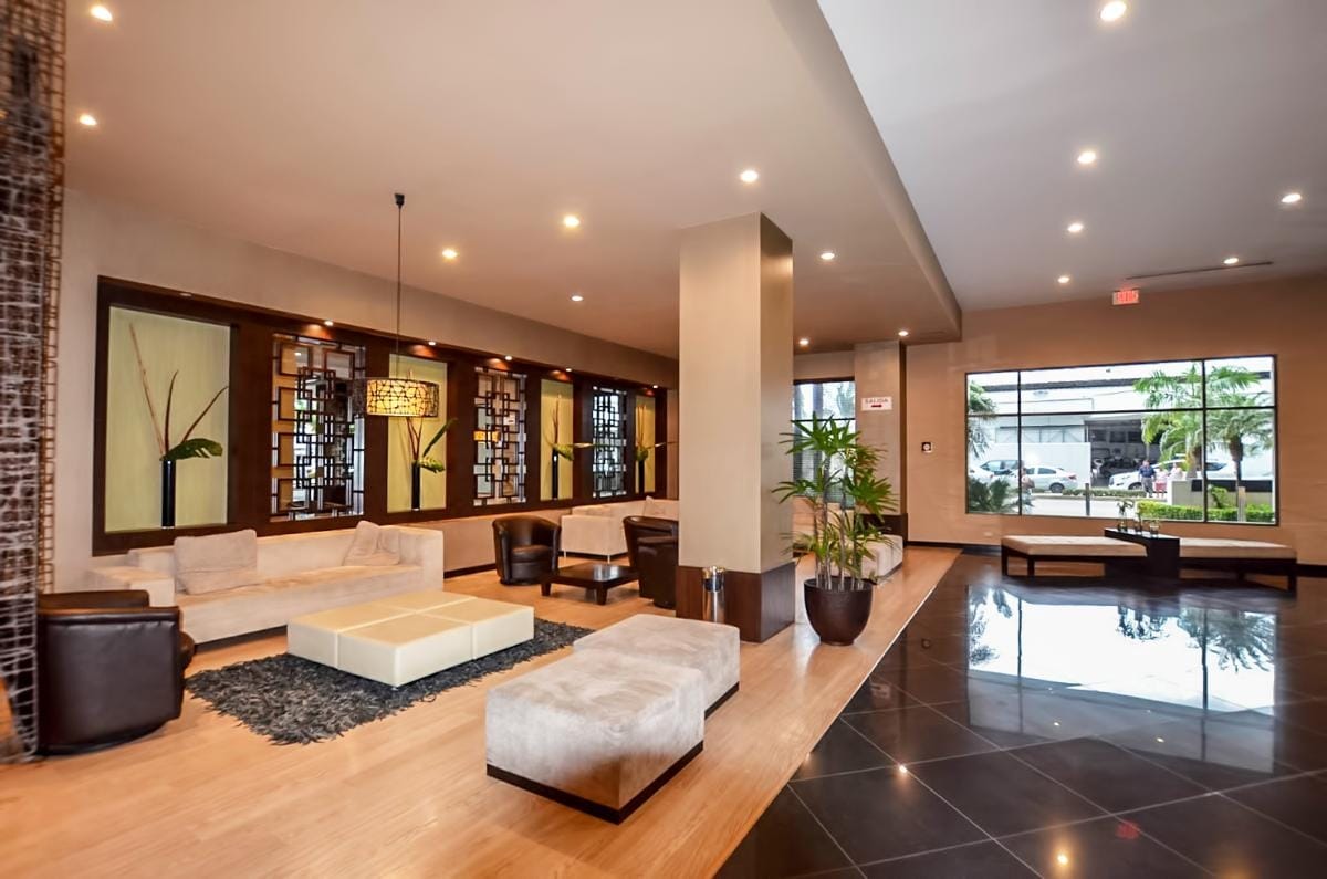 A spacious lobby area is highlighted by modern furnishings, including a low coffee table and comfortable seating. Large windows provide natural light and a view of the exterior, while decorative elements and plants add touches of style to the welcoming environment.