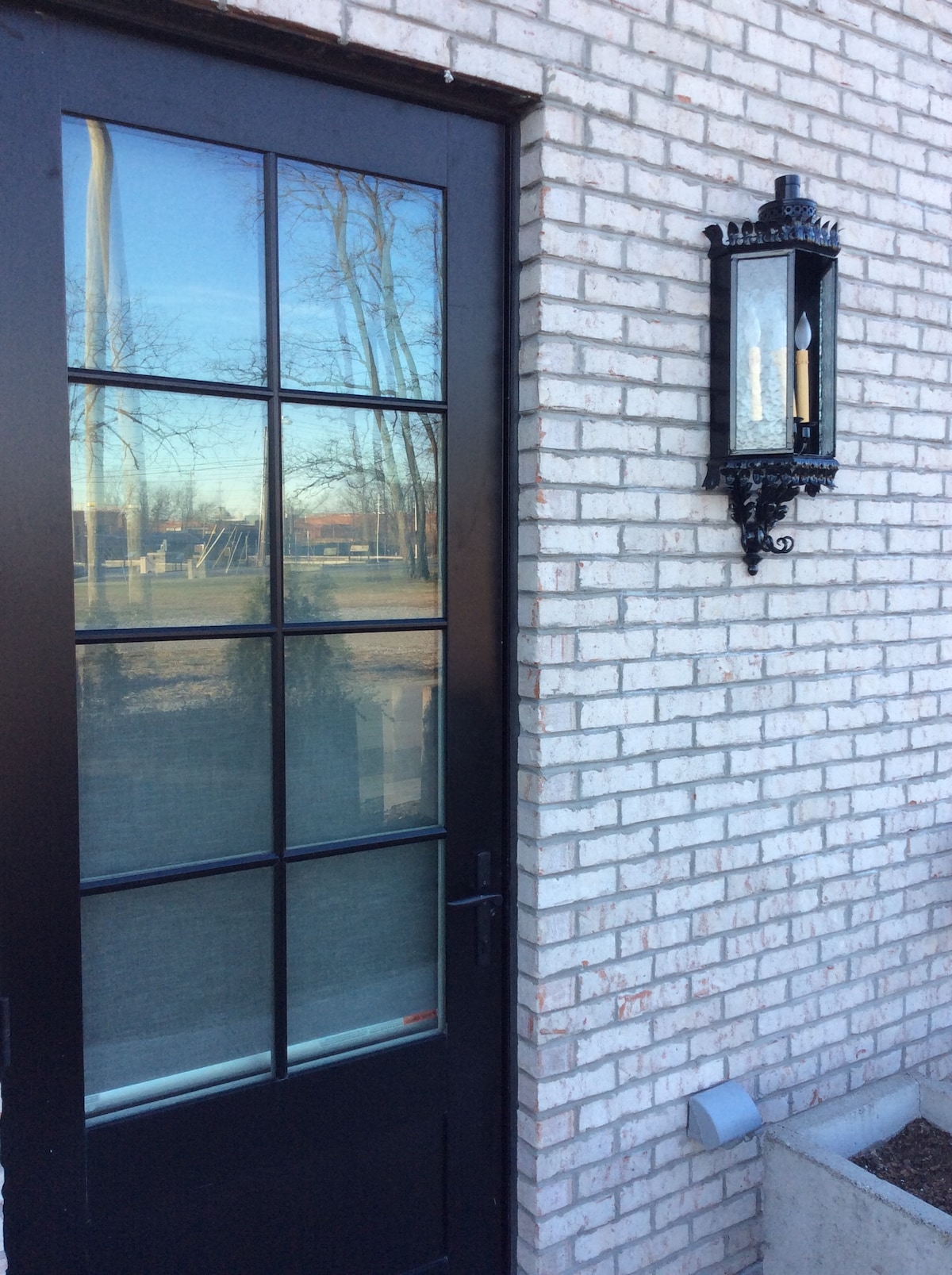 An entry door with large glass panes is framed by a white brick wall. A decorative lantern hangs beside the door, casting soft light. The reflection in the glass hints at nearby trees and open space, creating an inviting entrance to the suite.