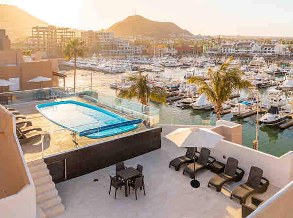 A rooftop pool glistens under the warm sun, with comfortable lounge chairs arranged nearby. The marina is visible in the background, lined with boats and framed by mountains, providing a serene setting for relaxation.