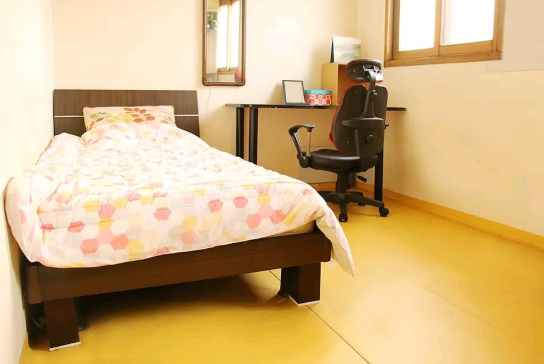 A simple bedroom features a spacious bed with a patterned blanket, complemented by a wooden frame. A desk with a chair is positioned next to a window, allowing natural light to brighten the area. The floor is finished in a light yellow hue, creating a warm environment.