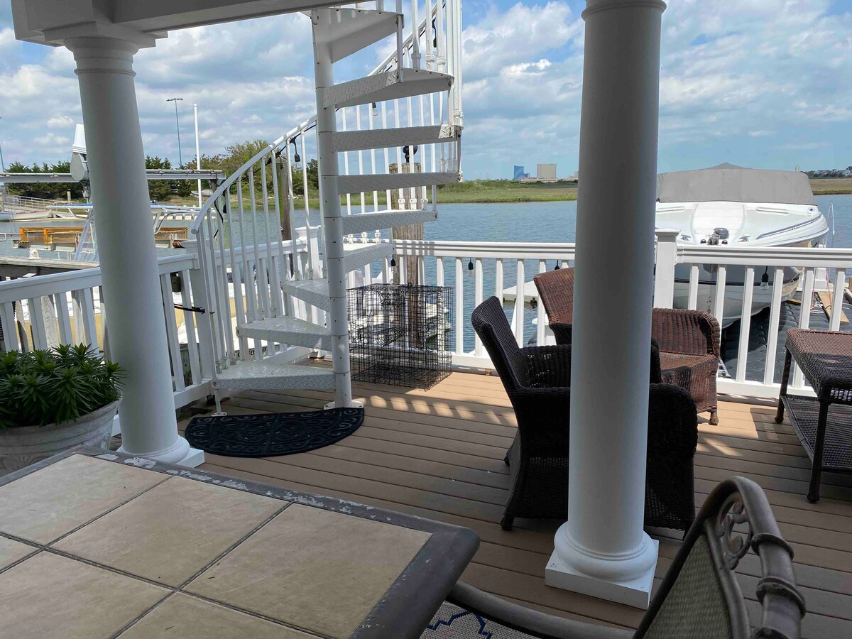 A spacious deck features a spiral staircase leading to an upper level, surrounded by seating areas that include wicker chairs and a table. The serene waterfront is visible beyond the railing, with boats docked nearby and a cloudy sky above.