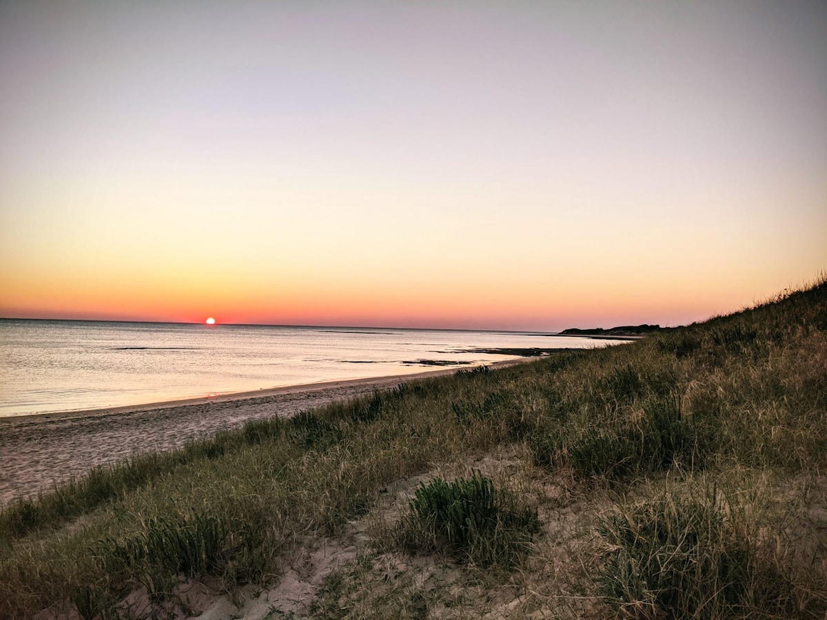 The image captures a serene beach at sunset, with gentle waves lapping at the shore. The sky transitions from soft orange to deep blue, mirroring the calm surface of the water. Grassy dunes are positioned along the sandy beach, contributing to the tranquil atmosphere.