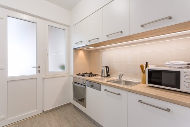 A modern kitchen space features sleek white cabinetry and stainless-steel appliances. A gas stove and sink are positioned on the countertop, with a microwave and kettle readily accessible. Natural light enters through a window beside the door, enhancing the clean and functional design.