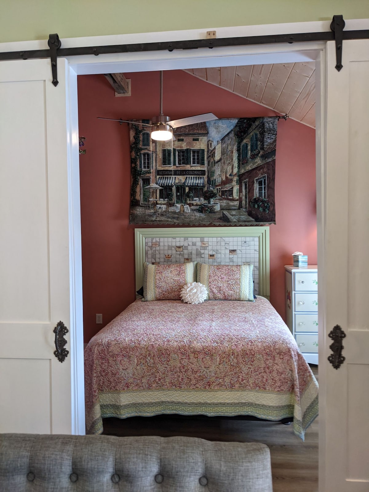 A cozy bedroom is framed by sliding barn doors, showcasing a spacious bed dressed in a floral quilt. Warm, peach-colored walls complement the intricate tapestry hanging above. A ceiling fan is positioned above, with a light fixture enhancing the bright atmosphere. A nightstand and dresser provide additional storage.
