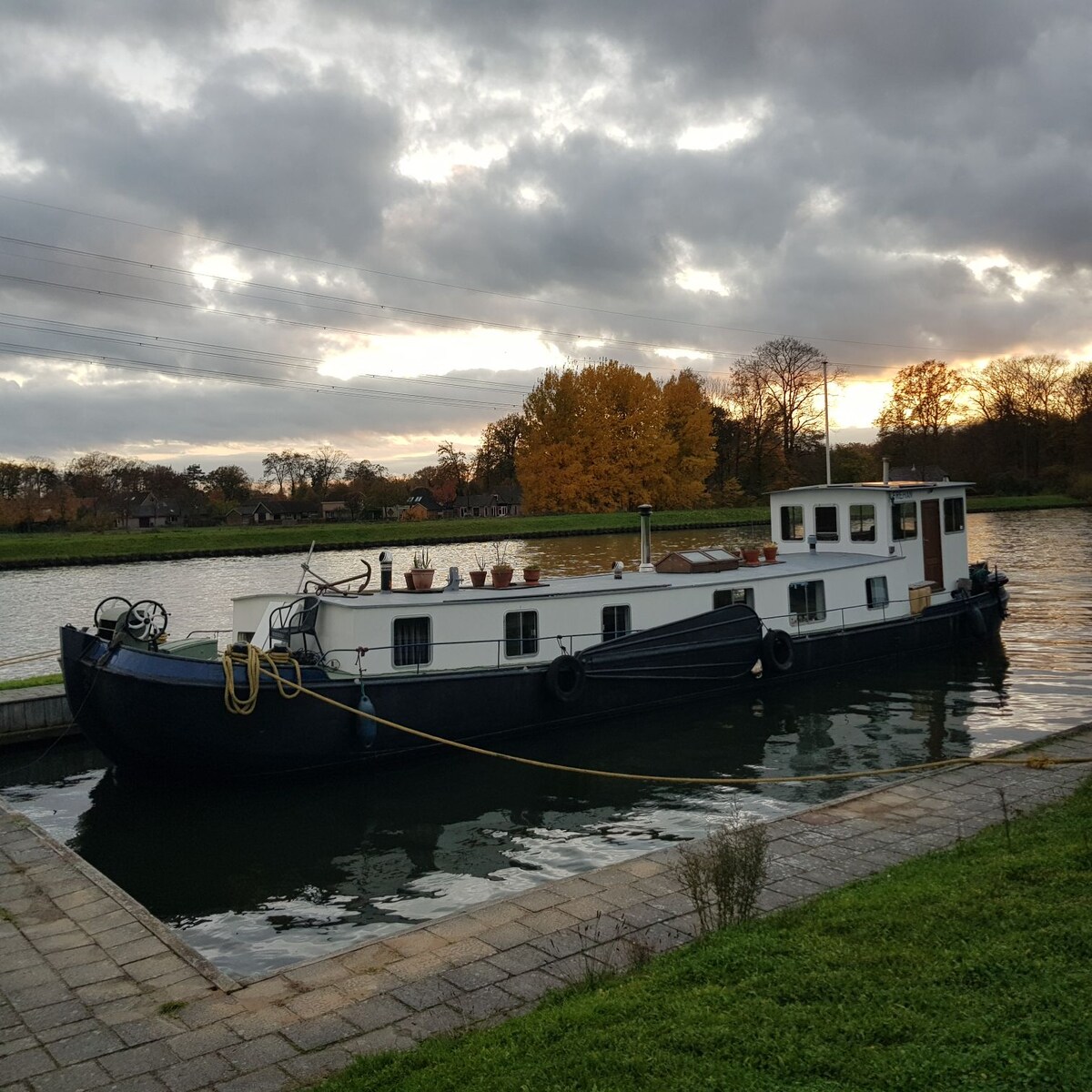 A vintage houseboat is docked along a serene waterway, surrounded by lush greenery and autumnal trees. The boat features a white exterior with several planters along the deck, reflecting a tranquil atmosphere as the sun sets in the background.