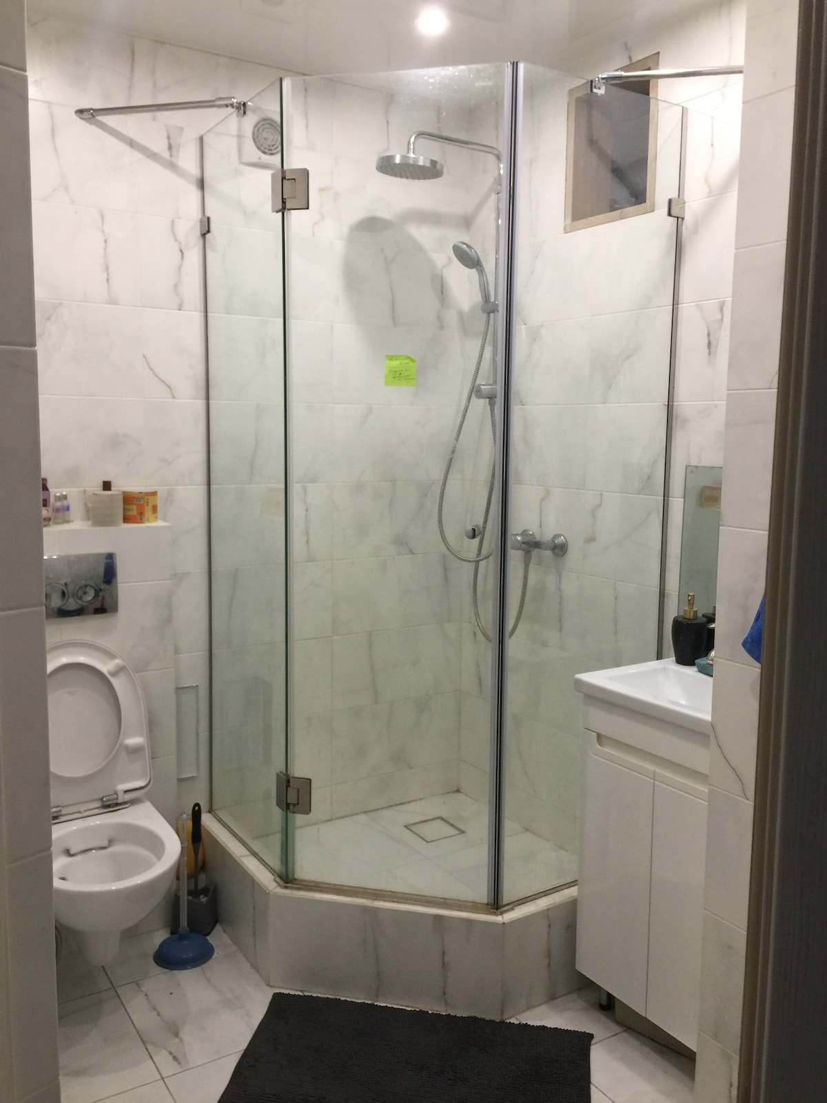 A modern bathroom is featured, showcasing a glass-enclosed shower with adjustable showerhead. A compact vanity with a sink is positioned beside the shower. Light tiles and a dark rug create contrast, while a small window allows natural light to enter the space.