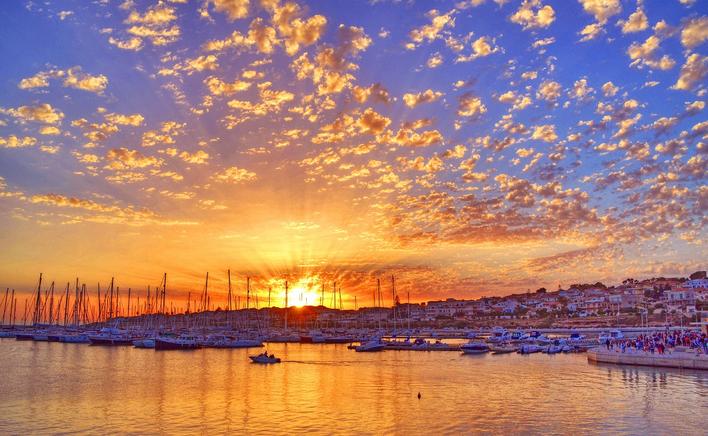 The image captures a vibrant sunset over a marina, with boats anchored in the calm waters. The sky is filled with colorful clouds transitioning from orange to blue, while the silhouetted masts of boats add depth to the scene. Gentle ripples reflect the hues of the sunset.