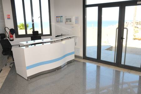A modern reception area features a sleek white desk with a light blue design. Large windows allow natural light to fill the space, offering a view of the ocean. Black chairs are positioned nearby, and polished flooring adds to the clean aesthetic.