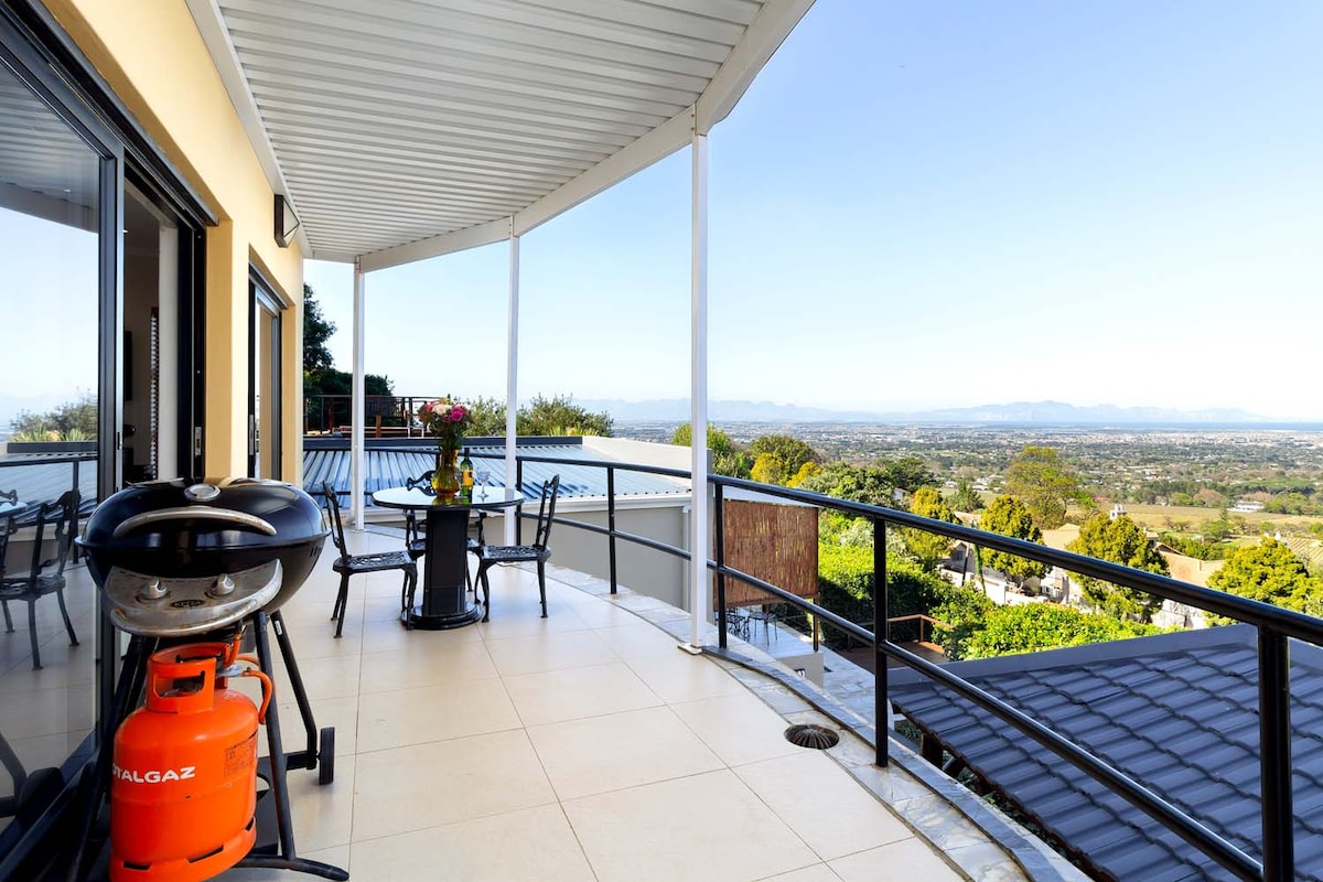 An outdoor terrace showcases a sleek barbecue and a round dining table with chairs, surrounded by expansive views of the Constantia Valley. The area is covered by a sloped roof, offering shade while allowing ample natural light to enter.