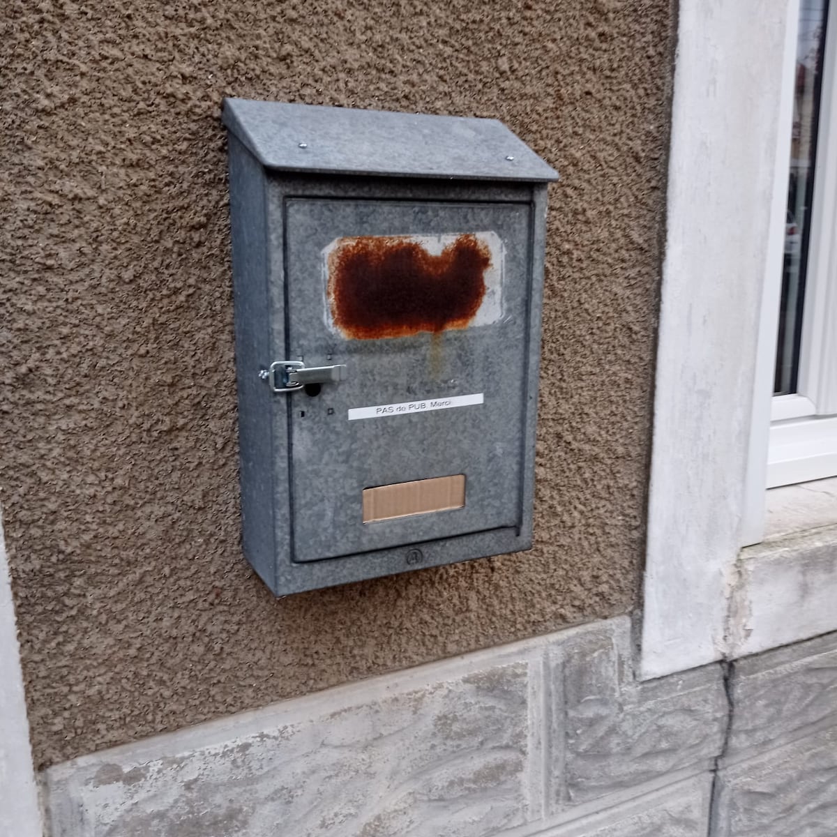 A wall-mounted mailbox is visible, featuring a weathered metallic exterior with signs of rust. A small lock is present, and a label indicating 'PASSE PAS' is affixed to the front. The mailbox is positioned against a textured wall, enhancing its rustic appearance.