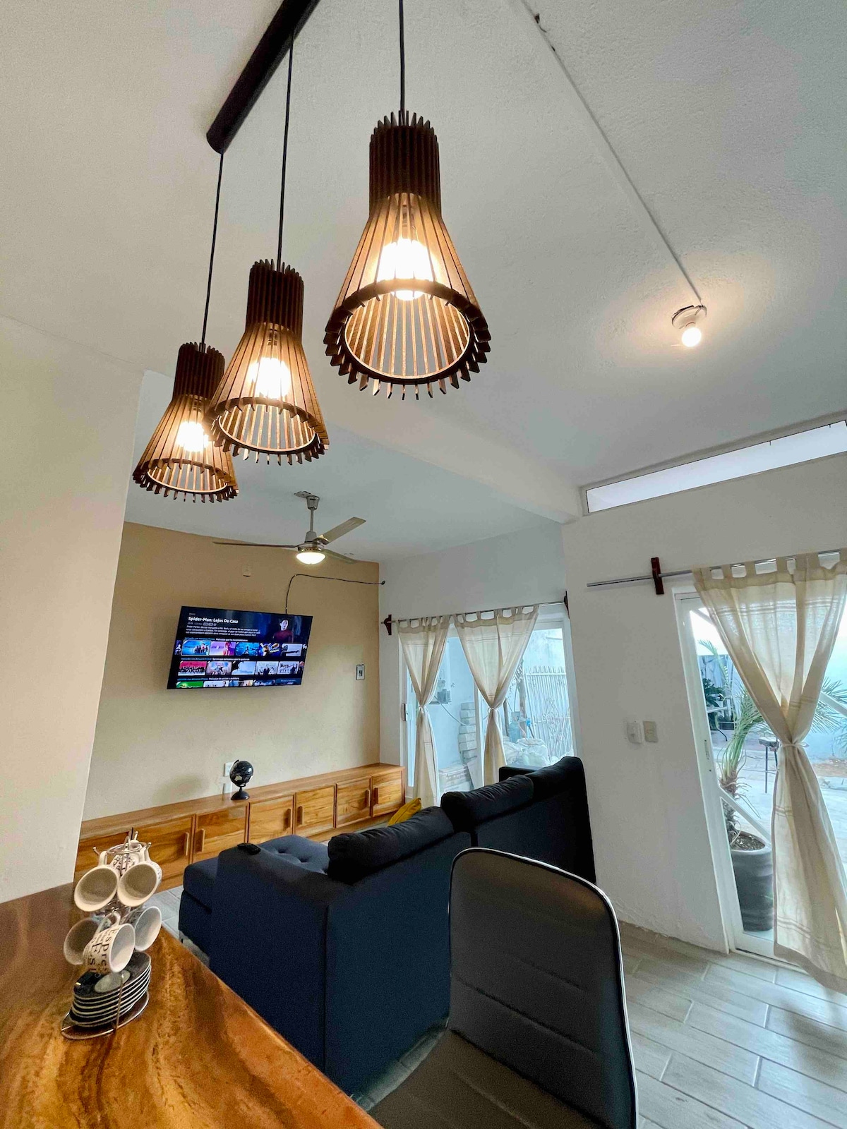 The living area features a comfortable sofa facing a wall-mounted smart TV. Three pendant lights hang from the ceiling, providing illumination. Floor-to-ceiling windows offer natural light, while doors lead to the outdoor space. A dining table and chairs are visible in the background.
