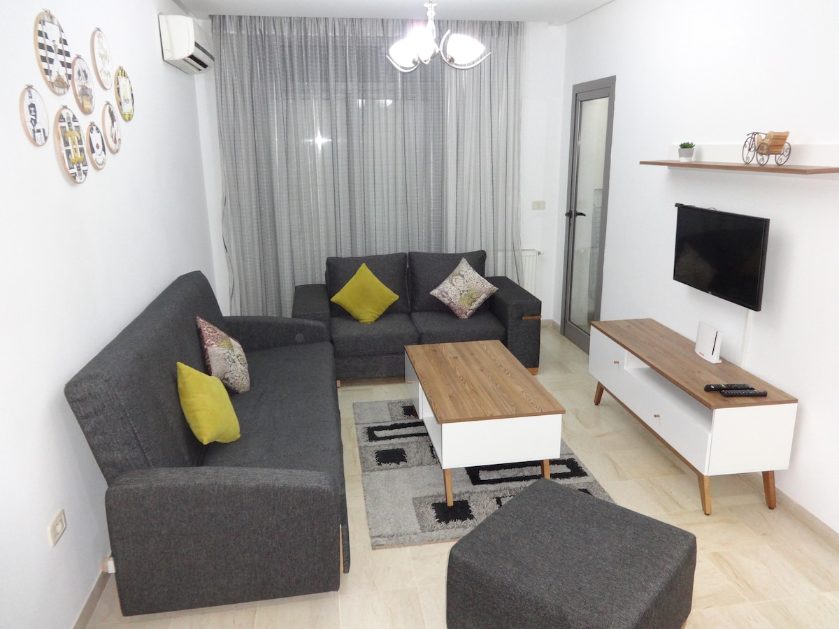 A spacious living area features a gray sectional sofa and a coordinating ottoman, with bright accent pillows adding color. A wooden coffee table is centered on a patterned rug, complemented by a white media console under a flat-screen television. Sheer curtains allow natural light to filter in.