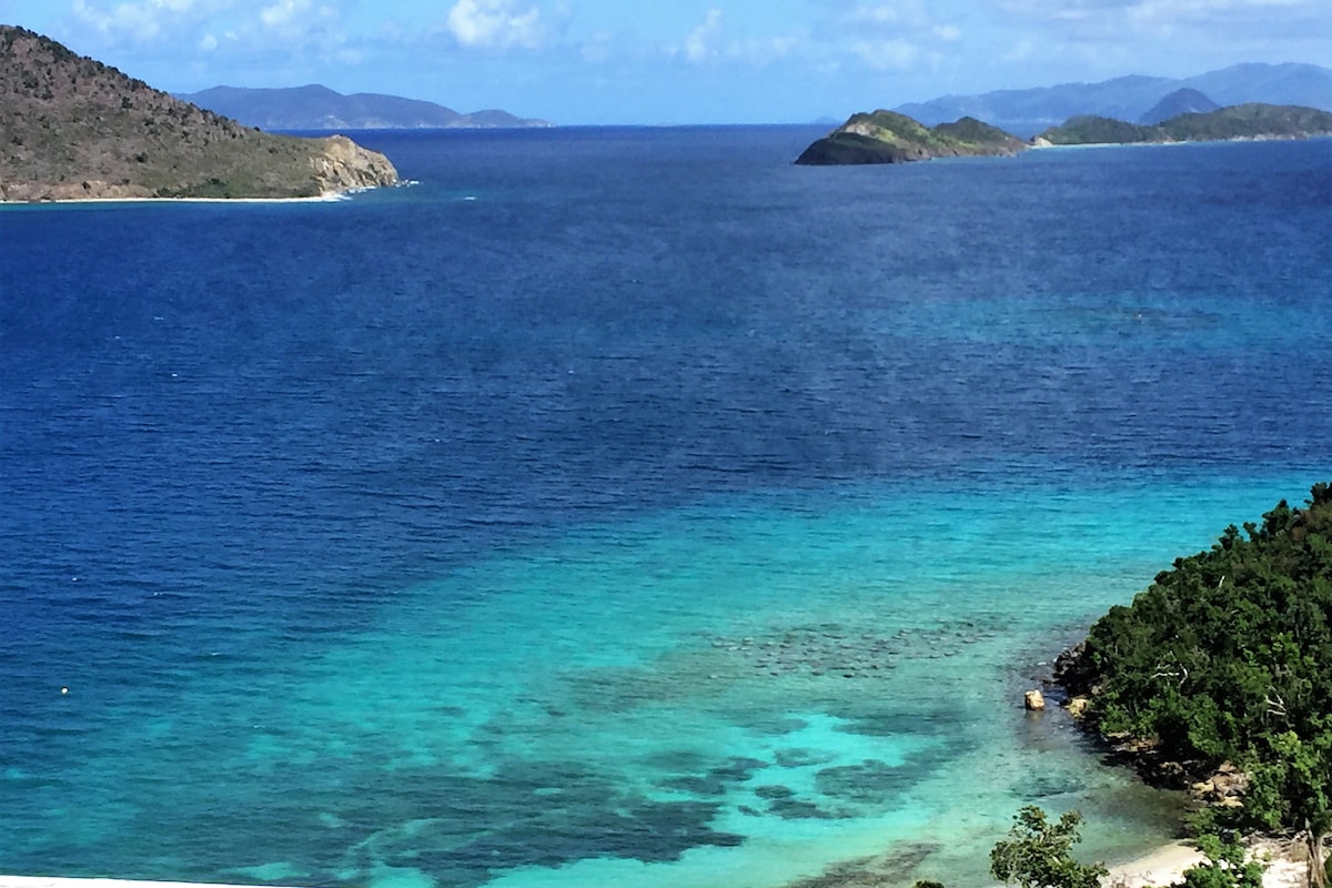 A serene view of the ocean is captured, showcasing varying shades of blue and turquoise waters. Islands are visible in the distance, framed by lush greenery along the coastline. The clear sky enhances the tranquil atmosphere, inviting relaxation and exploration.