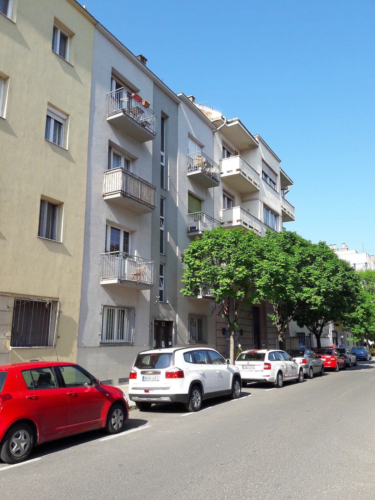 A colorful building is shown with multiple balconies and varying façade colors. Trees provide shade along the street, where parked cars are lined up on both sides. The clear blue sky adds to the bright appearance of the neighborhood.