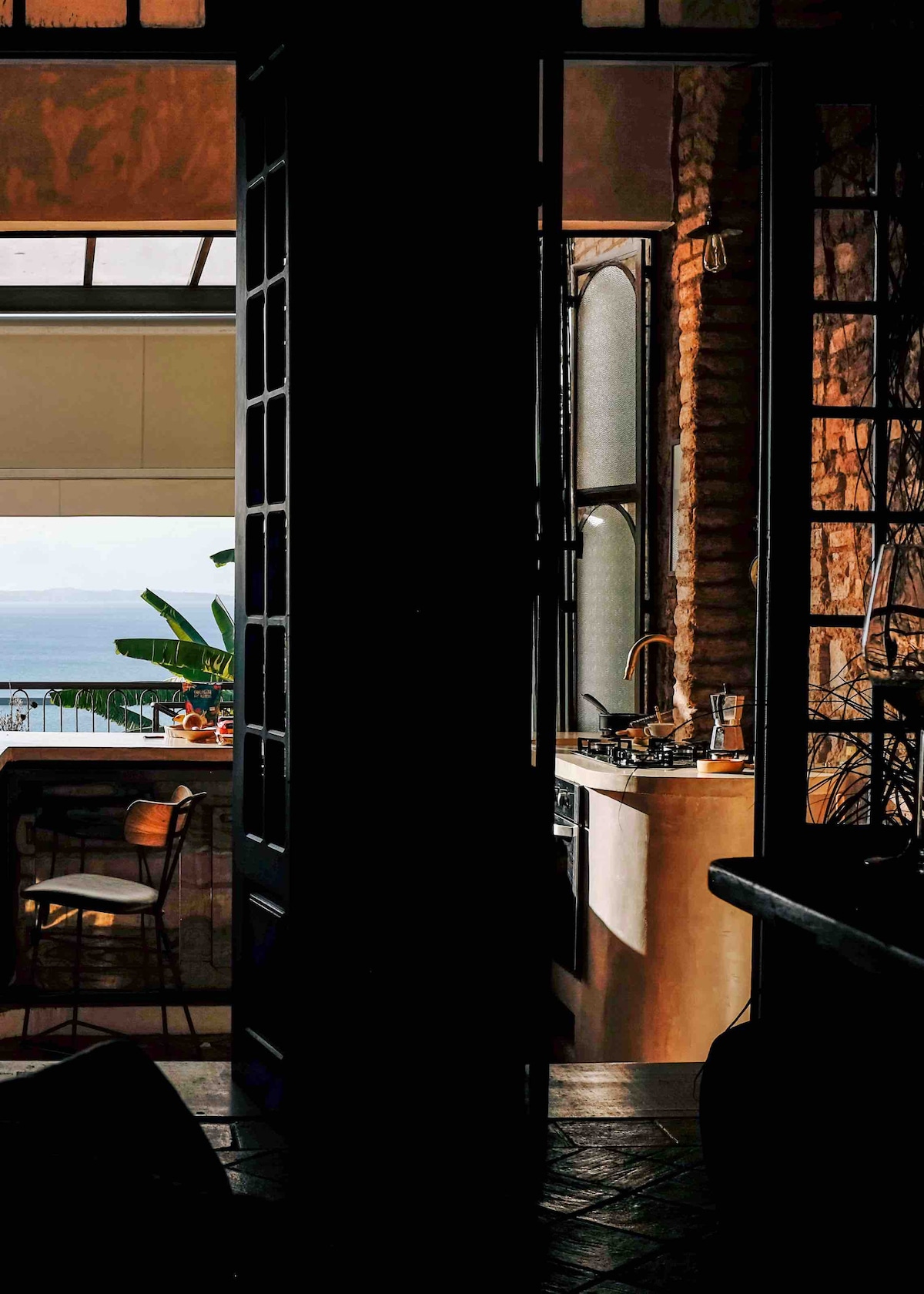 An inviting kitchen space is framed by open doors, revealing a view of the sea. Natural light filters through large windows, highlighting modern appliances and a cozy dining area with a wooden bar. Green plants can be seen beyond the kitchen, contributing to the serene atmosphere.
