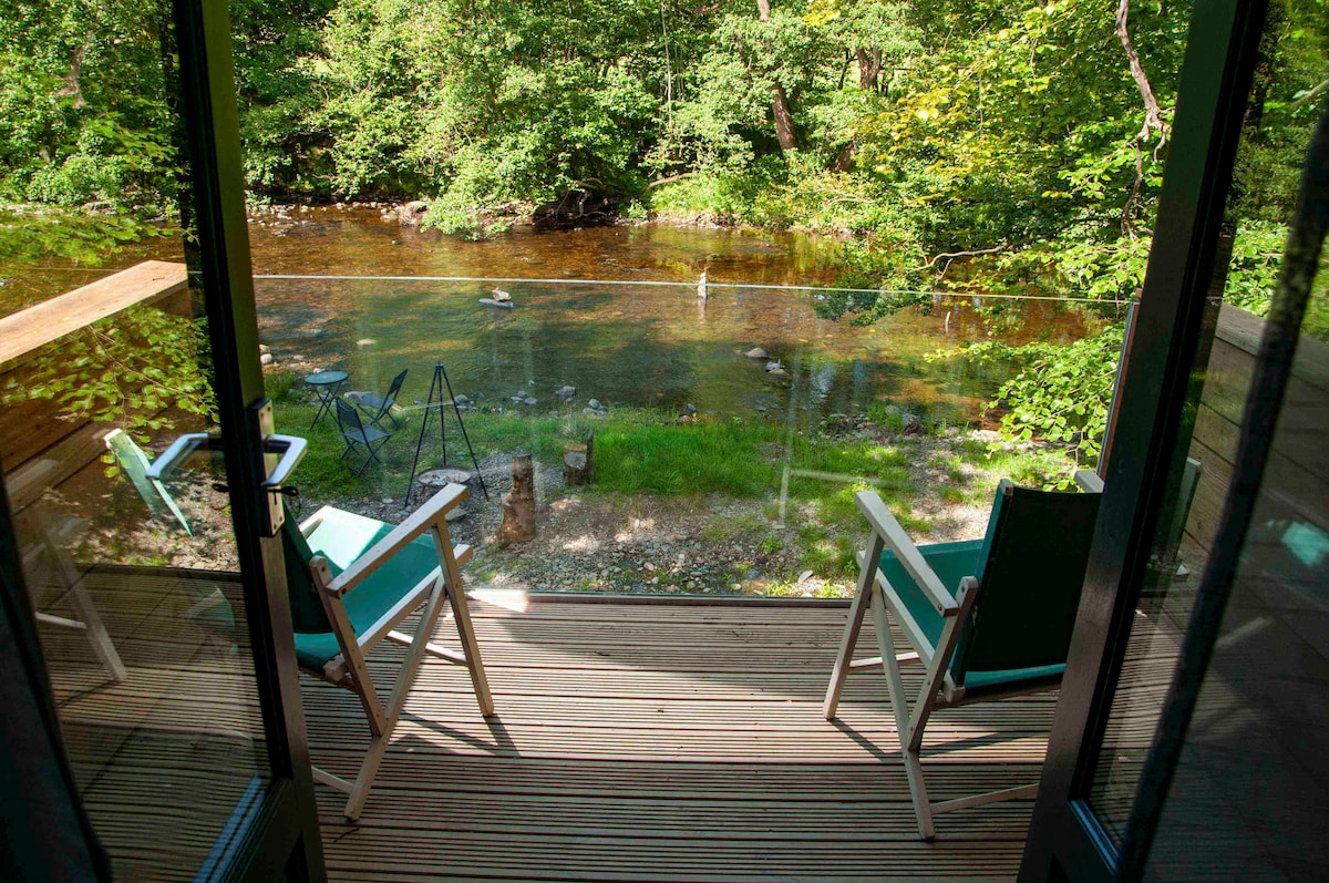A wooden terrace offers two light green chairs facing the serene River Vyrnwy. Surrounding foliage creates a natural setting, while the calm water reflects surrounding greenery. The view captures a peaceful outdoor space ideal for relaxation and birdwatching.