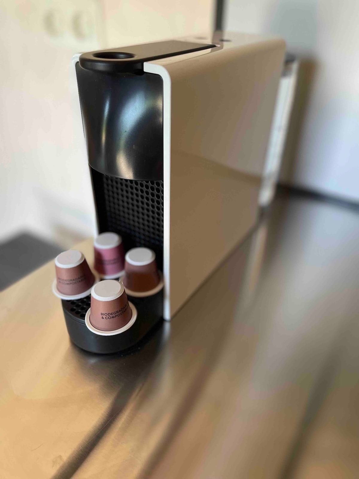 A coffee machine is positioned on a metallic countertop, featuring a sleek white and black design. A holder with four coffee pods rests at the base, showcasing a mix of colors. Natural light subtly illuminates the scene.