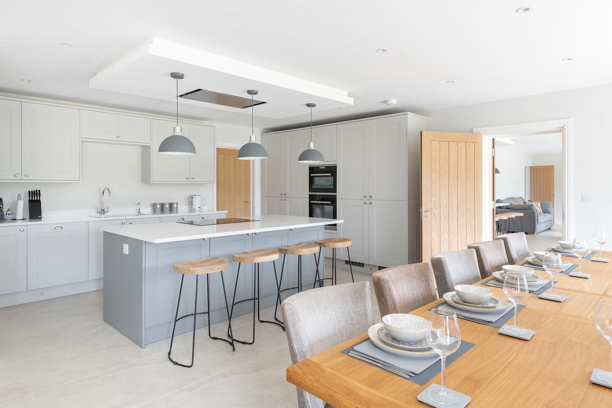 A modern kitchen is presented with sleek cabinetry and an island bar featuring high stools. A dining table accommodates multiple settings with elegant dishware and glassware. Natural light illuminates the space, enhancing the clean lines and spaciousness of the area.