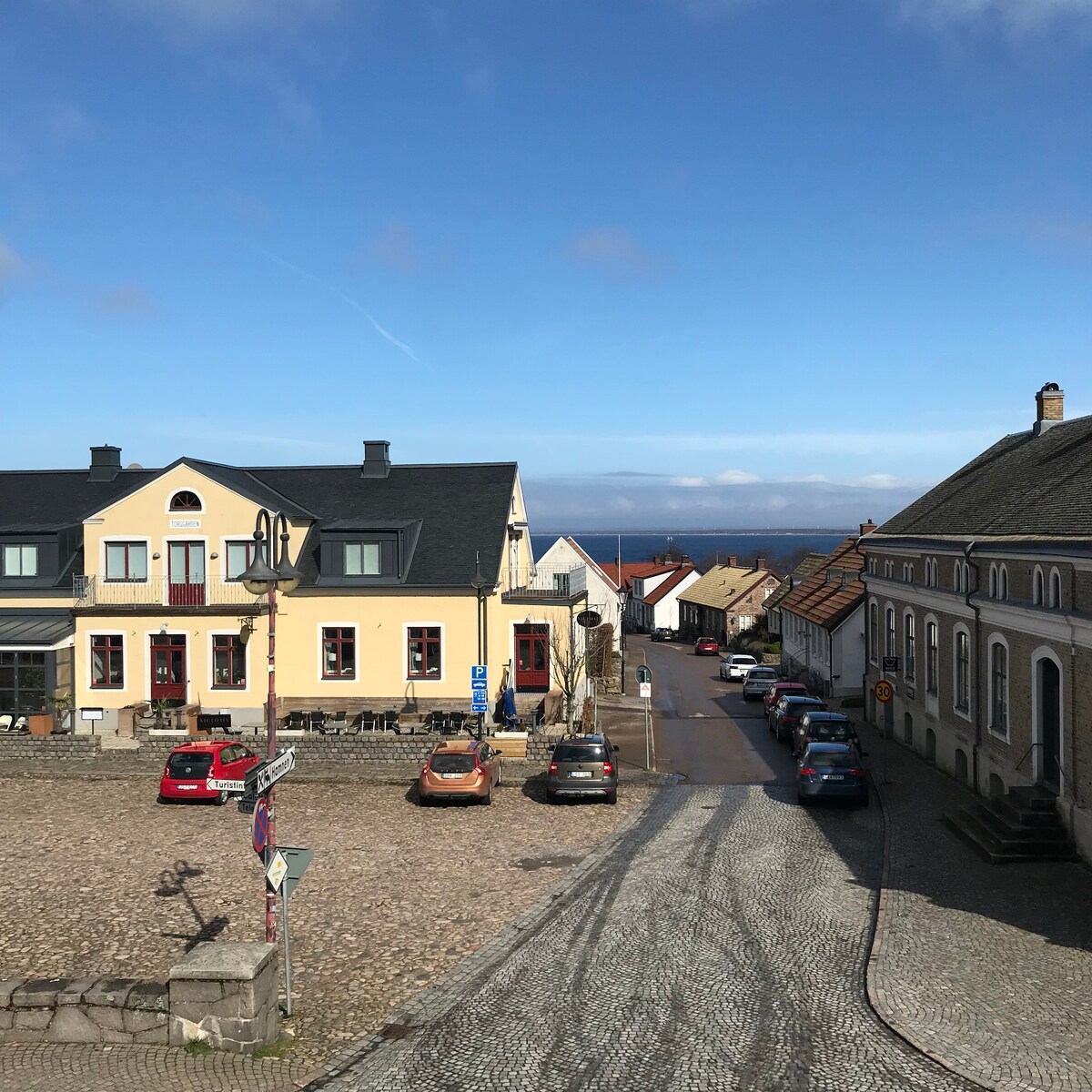 A view of a cobblestone street is presented, lined with charming buildings featuring varying architectural styles. Parked cars are visible along the side, while the distant horizon showcases the sea under a clear blue sky.