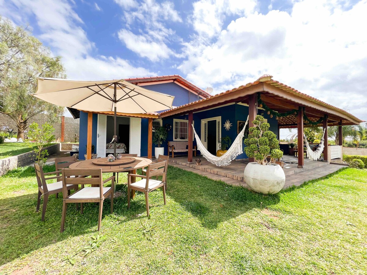 An outdoor seating area features a round table surrounded by four chairs, set under a large umbrella. A spacious patio is decorated with hammocks and greenery, providing a peaceful environment. The house is painted in blue, with a terracotta roof complementing the landscape.