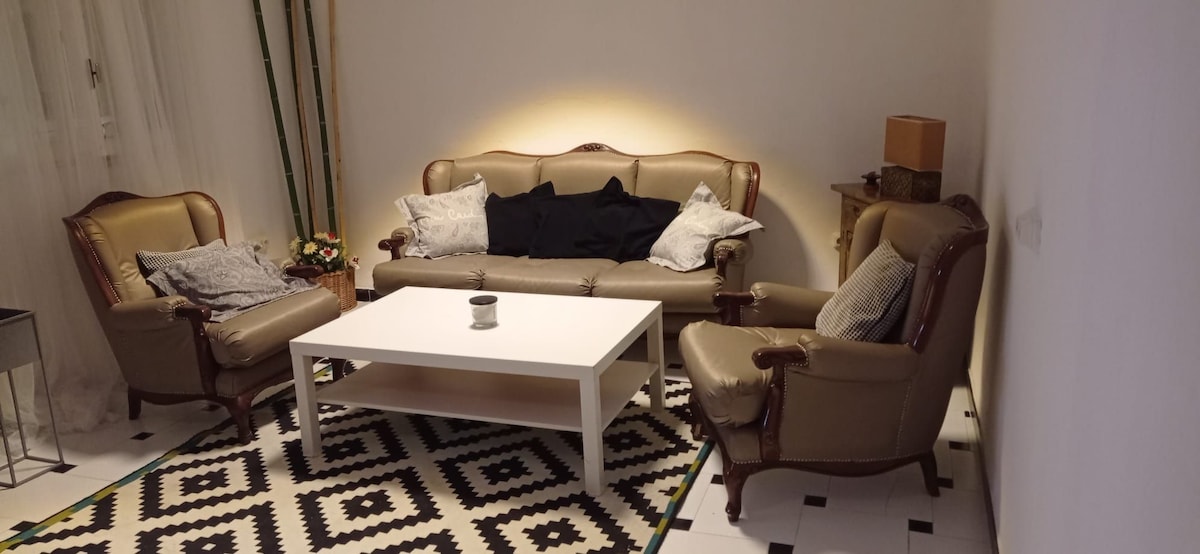 A living area is showcased, featuring a light-colored sofa and two matching armchairs, all arranged around a low, white coffee table. A patterned rug lies underneath, and soft lighting highlights the seating area, enhancing the inviting atmosphere.