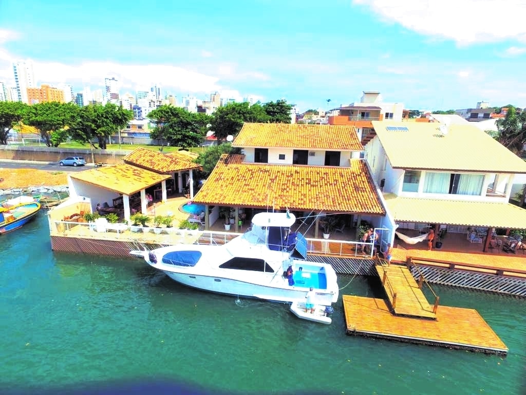 A waterfront property is showcased, featuring a house with a tiled roof next to a moored boat. A wooden deck extends into the water, with lush greenery and urban buildings visible in the background, enhancing the scenic canal-side location.