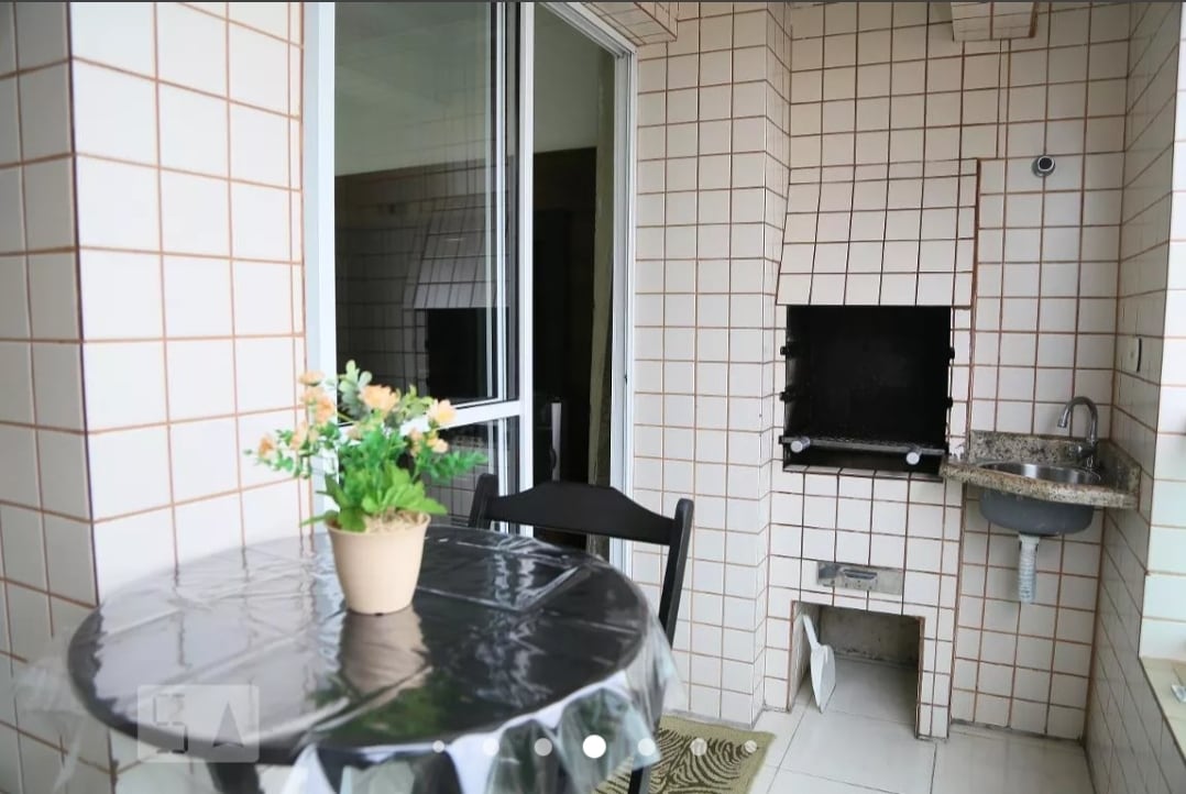 A balcony space features a round table covered with a silver tablecloth and a potted plant. A built-in barbecue grill is positioned against the tiled wall, alongside a small sink area. Natural light illuminates the tiled surfaces.