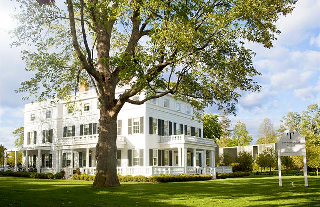 A charming three-story white building is framed by a large, leafy tree, creating a welcoming façade. Black shutters adorn the windows, while the expansive front porch provides a space for relaxation. Lush green grass surrounds the property, enhancing the inviting ambiance.