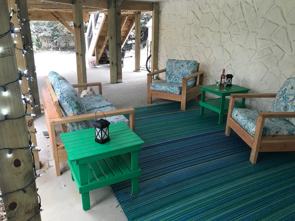 A covered patio area features comfortable seating arranged around green side tables. The space is brightened by decorative lighting, while a large, stylish rug provides a cozy foundation. Natural textures and colours create a welcoming environment for relaxation.