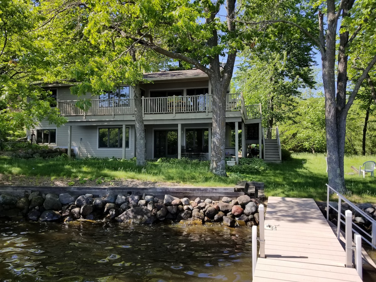 A lakefront property is framed by lush green trees, showcasing a multi-level structure with a spacious deck. The lower level features large windows, while a private dock extends over the serene water, complemented by a stone shoreline.