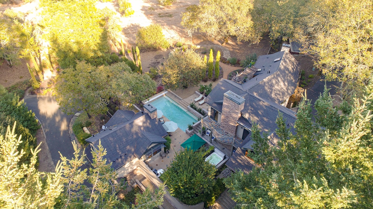 An aerial view captures the spacious property, showcasing a large pool surrounded by lounge areas and gardens. The two-story main house features a combination of dark roofing and natural surroundings, while trees and landscaped areas provide greenery around the retreat.