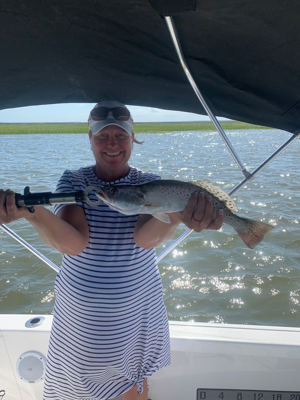 An individual is holding a freshly caught fish while standing on a boat under a shaded canopy. The water is visible in the background, reflecting sunlight. The person is wearing a striped dress, and the focus is on the vibrant fish being displayed.