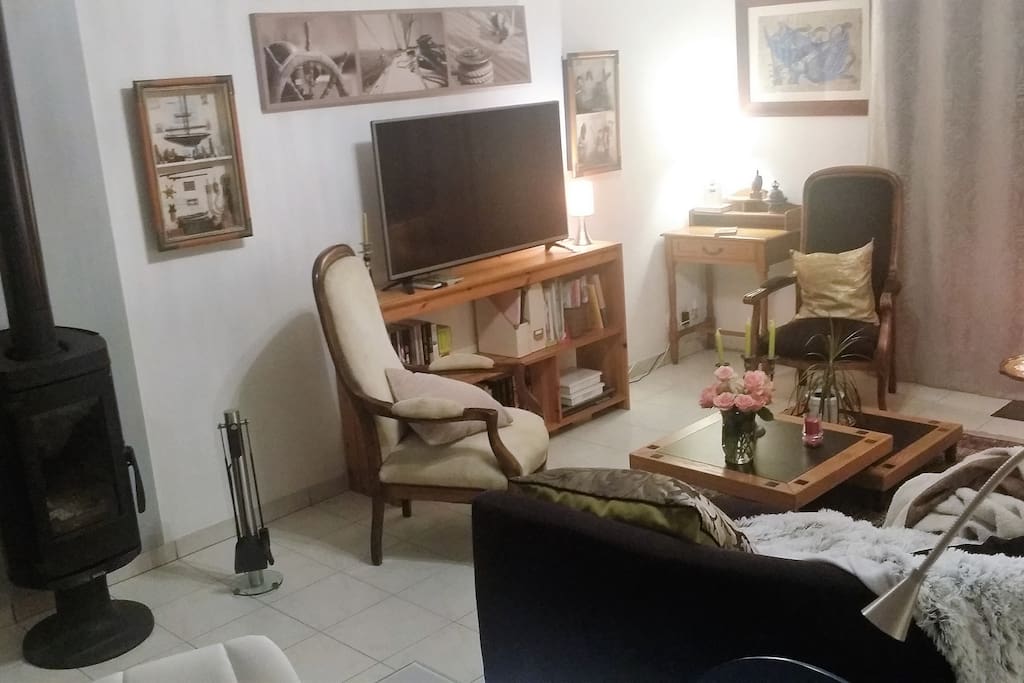 le salon avec le poele à bois, le canapé d'angle le grand écran tv et des romans à disposition.