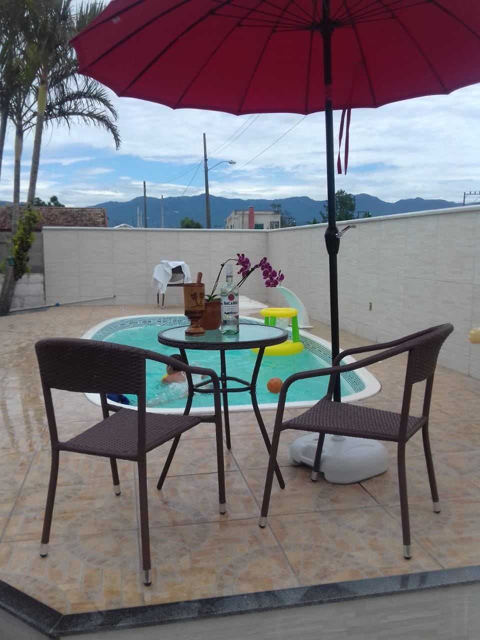 A small pool is visible, surrounded by a tiled area with two wicker chairs and a table. An umbrella provides shade over the seating. In the background, mountains are faintly seen, contributing to a serene outdoor setting.