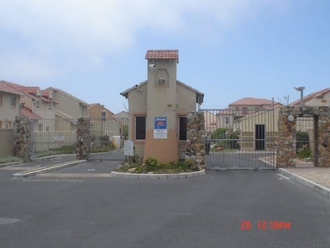 A gated entrance is shown, featuring stone columns flanking a tall security gate. A small building with a peaked roof stands beside the gate, housing a security system. Clear signage indicating the community's name is visible. Surrounding buildings and landscaped areas can be seen in the background.