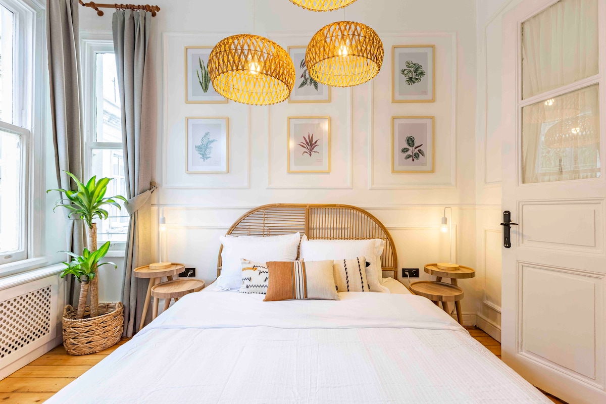 A bright bedroom features a spacious bed with crisp white linens, accompanied by two small side tables. Above the bed, decorative wall art displays botanical prints. Two woven pendant lights provide soft illumination, and a green plant adds a touch of nature to the serene atmosphere.
