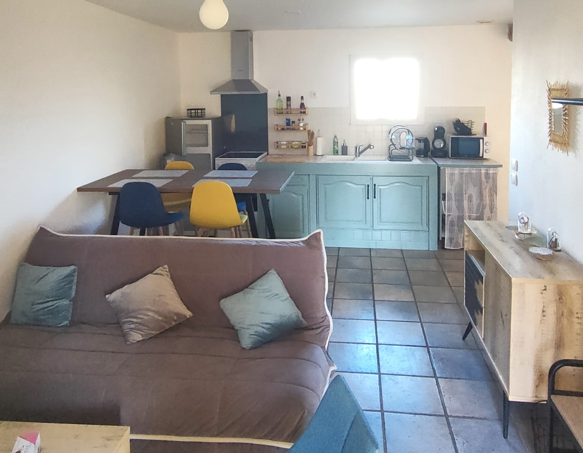 An open living area features a spacious sofa adorned with cushions, a dining table with colorful chairs, and a modern kitchen equipped with essential appliances. Natural light floods the space through a window above the sink, illuminating the tiled floor and the light-colored cabinetry.
