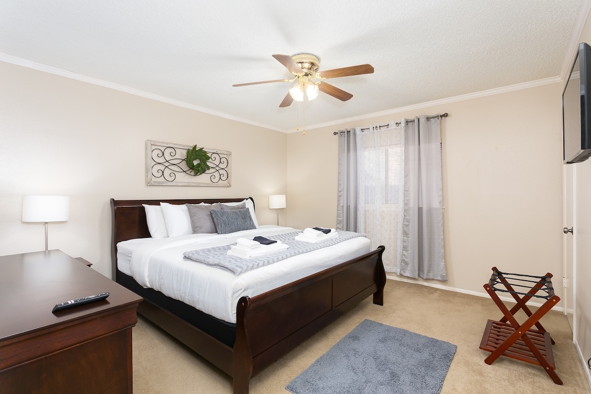 A tranquil bedroom features a spacious bed adorned with crisp linens and decorative pillows. Natural light enters through sheer curtains, illuminating the room. A ceiling fan provides airflow, while a television is mounted on the wall. A dresser and a folding luggage rack offer added convenience.
