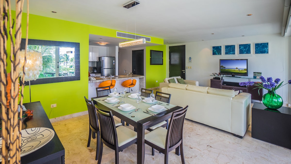 A modern living space features vibrant green walls that complement the sleek black dining table set for six. A spacious sofa faces a wall-mounted television, while a kitchen area is visible in the background. Artistic decor and a colorful vase add a touch of elegance.