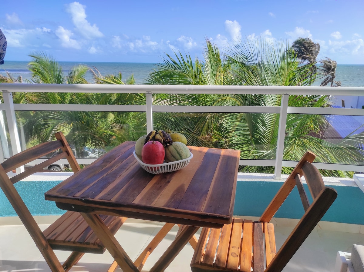 A balcony is featured with a wooden table and two folding chairs, overlooking the ocean. A bowl of assorted fruits is centered on the table. Lush palm trees and a clear blue sky with scattered clouds are visible in the background.