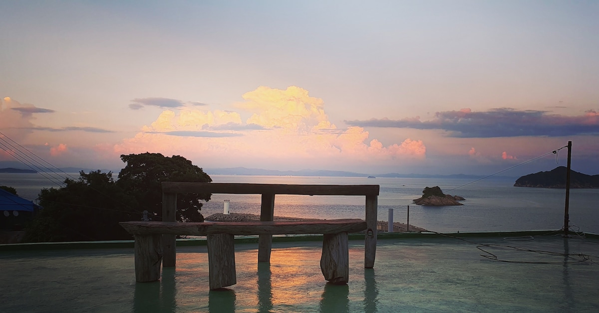 A wooden bench is positioned on a green terrace overlooking a calm sea. The horizon features soft pastel clouds illuminated by the setting sun, while an island is visible in the distance, contributing to the tranquil setting.