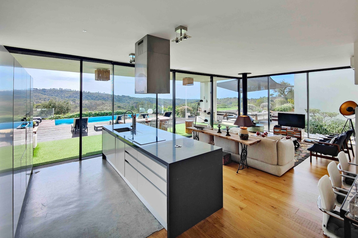 The modern kitchen transitions into an open living area, featuring large windows that offer views of the pool and valley beyond. A central island with seating provides a gathering space, while natural light highlights the sleek design and neutral color palette.
