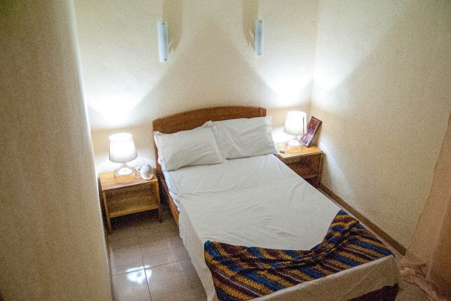 A bright sleeping area features a spacious bed with crisp white linens. Two bedside tables are positioned on either side, each adorned with a soft lamp. Natural light filters in softly, creating a calm and comfortable environment.