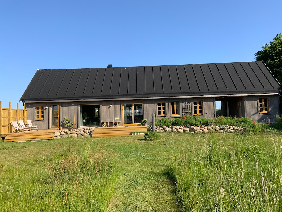 A modern house with a black roof and wooden exterior is framed by lush green grass. Large windows provide views of the surrounding landscape, while a spacious deck features lounge chairs and is bordered by decorative stones and plants.