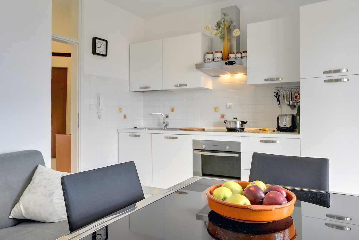 A bright kitchen area is presented with white cabinetry and a modern design. A glass-top dining table with a bowl of assorted apples and pears is displayed in the foreground. Essential appliances, including an oven, stove, and refrigerator, are conveniently arranged, providing a functional cooking space.