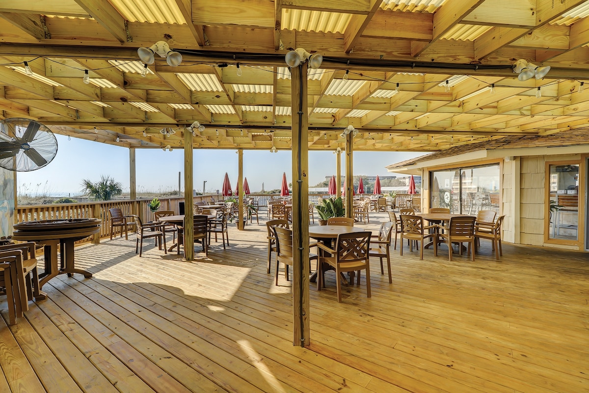An open-air dining area is featured under a wooden pergola, providing ample shade. Multiple wooden tables and chairs are arranged throughout the spacious deck. Bright red umbrellas offer additional shade around the seating areas, with palm trees visible in the background against a clear sky.