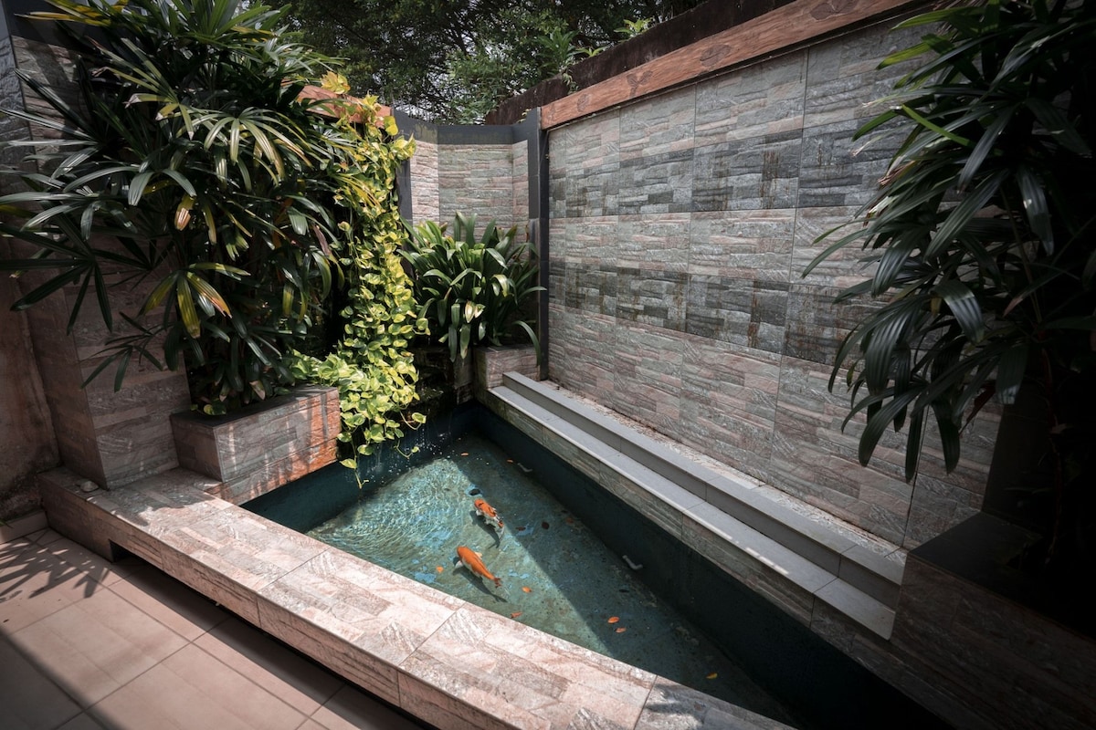 A serene outdoor pond is surrounded by lush green foliage and textured stone walls. The clear water reflects vibrant orange koi fish swimming, while stone seating edges the pond, providing a tranquil space for relaxation.